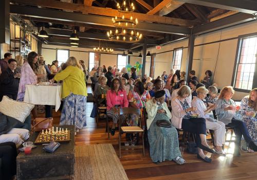 Spacious event room with a crowd, wooden beams, and chandeliers.