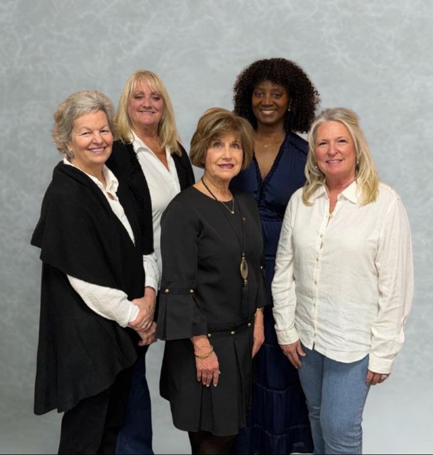 Five women standing together, smiling in front of a gray backdrop.