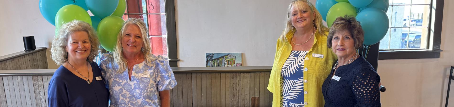 Four women smiling around a decorated cake with green and blue balloons in the background.
