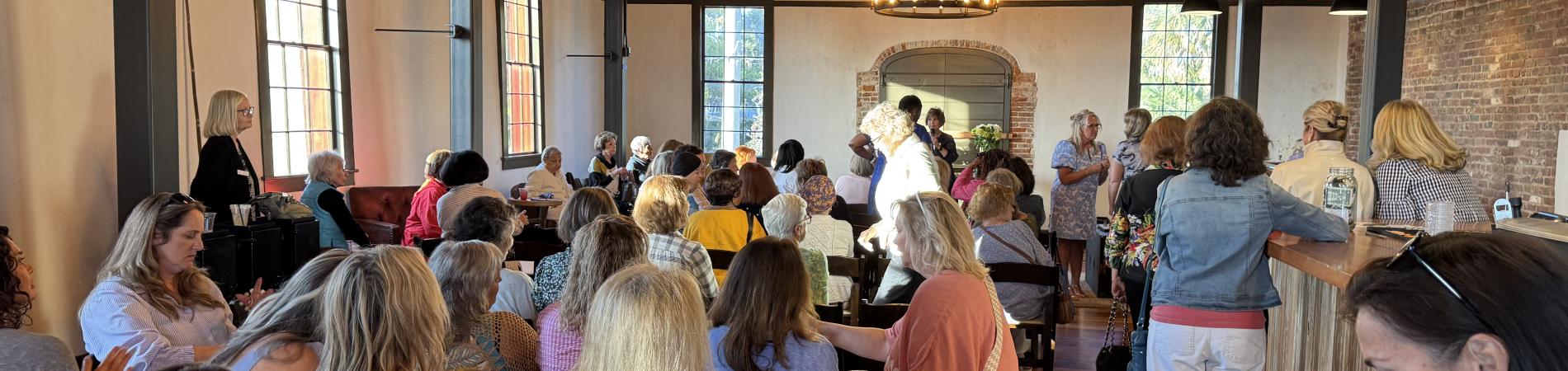 Crowded room with people sitting and standing, warm lighting, large chandeliers above.